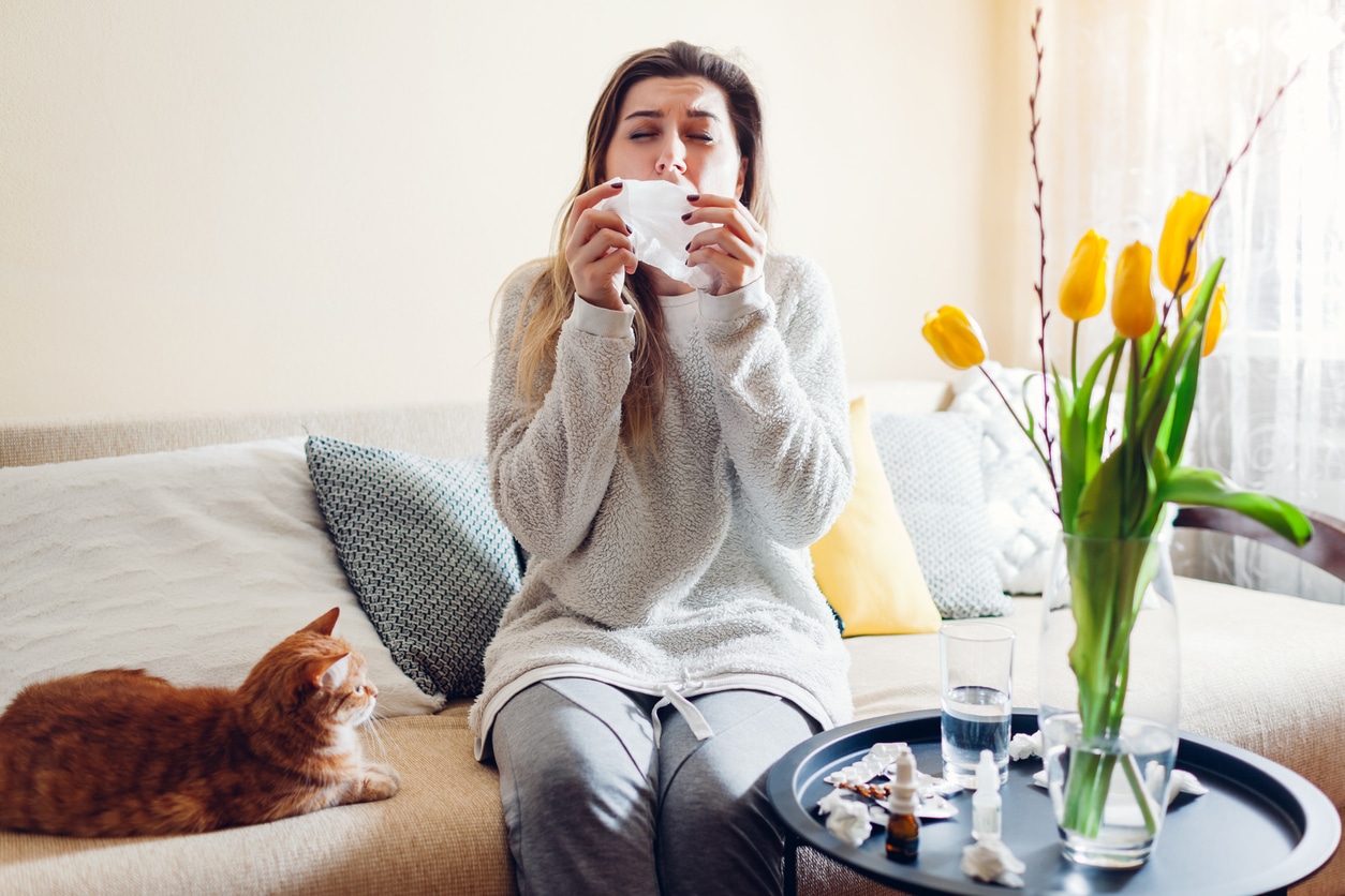 Woman sitting next to a cat in her home about to sneeze into a tissue.